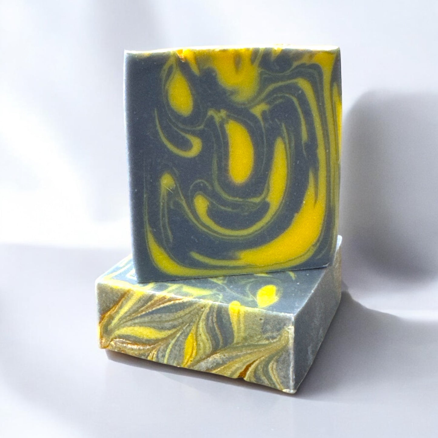 A bar of Dandy Moon Soapery's Astro mens Soap with a bold swirl design in dark navy blue and bright yellow. The bar sits atop another bar, to show the gold swirl design of the bar. Both bars are sitting on a white surface, against a white background. The contrasting colors and intricate marbling evoke a sense of energy and sophistication, making this handcrafted soap a luxurious choice.