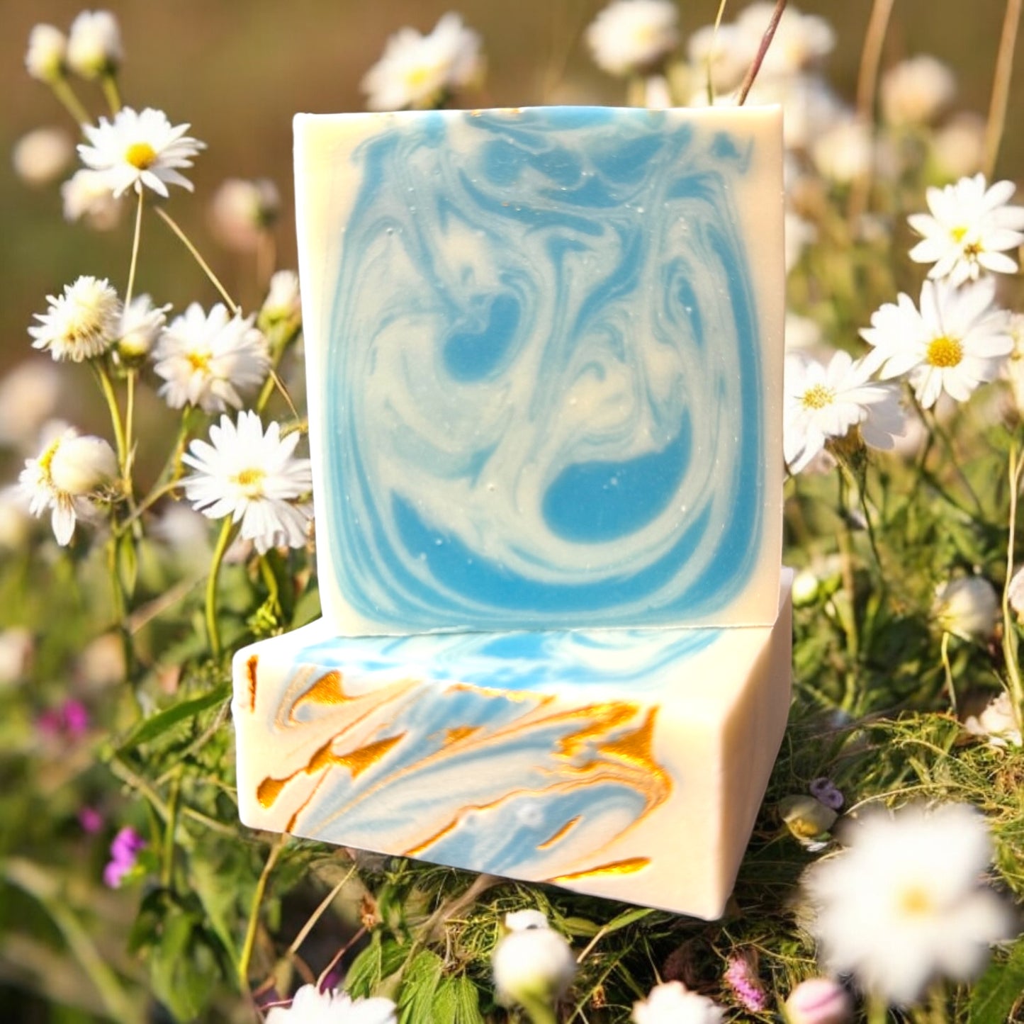 a bar of artisan soap with a vibrant swirl design in blue and white, set against a background of blooming daisies. The soap has a top design of swirls of blue and white and a splash of gold. The intricate marbling and bright colors capture a sense of tranquility and natural beauty, perfect for a refreshing and uplifting self care experience.