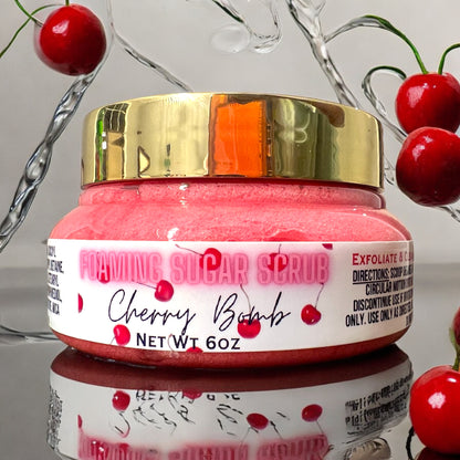 a jar of foaming sugar scrub, the jar is closed and the top of the jar lid is gold. it sits on a reflective surface and water is splashing around the jar. there are cherries falling all around the jar and sitting on the reflective surface. 