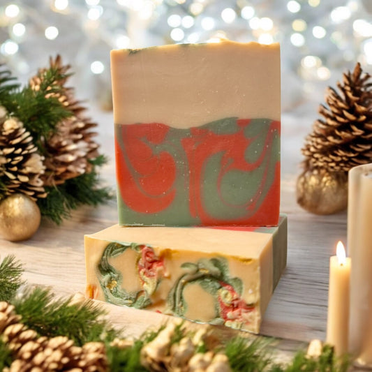 A bar of Dandy Moon Soapery's Noel soap is sitting on top of another bar that is laying flat, to showcase the top design with swirls of color that are Christmas wreaths, and displayed in a festive holiday setting with pinecones, evergreen branches, and softly glowing candles. The soap features a warm cream top layer with a bottom layer having swirls of red and green. The intricate marbling design adds a touch of elegance, while the Holiday themed decor around it captures the spirit of the season. 