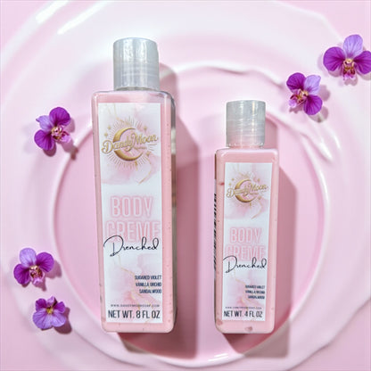 Two bottles of dandy moon body creme lotion in the scent drenched lay flat on a light pink rippled liquid surface surrounded by purple orchids. the bottles are clear and the lotion inside is a soft pink. 