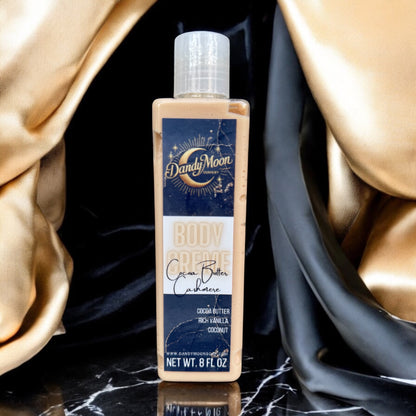 a bottle of body creme lotion in the scent cocoa butter cashmere stands on a black marble surface, against a background of black and gold satin fabric drapes