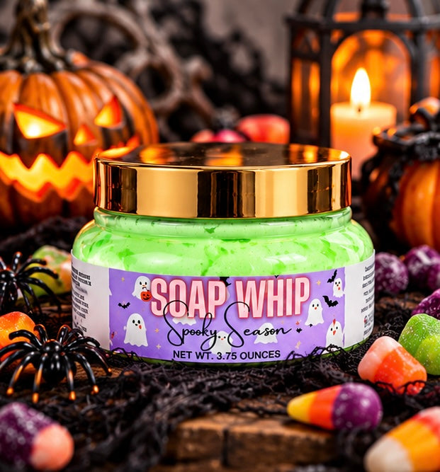 A jar of Halloween-themed 'Soap Whip' body soap with bright green soap Whip inside, a shiny gold lid, and a purple label with tiny white ghosts and reads 'SOAP WHIP in the scent 'spooky season'. The jar is styled on a black net with candy, spiderwebs, and jack-o'-lanterns in the background.