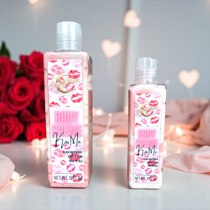 Two bottles of Body Creme Lotion in the scent kiss me with pink, the lotion bottles are clear and the lotion inside is white and reddish pink. The bottles stand on a white surface and in the blurred background is twinkle lights, pink satin fabric, and a dozen red roses bouquet.  