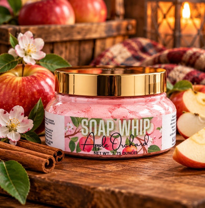 A jar of 'Soap Whip'  in the scent Apple Orchard sits on a rustic wooden table surrounded by fresh red apples, white blossoms, cinnamon sticks, and a warm, cozy background with a lantern and plaid blanket.