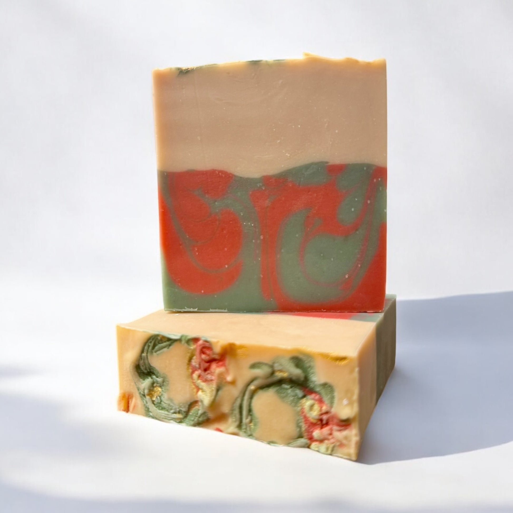 A bar of Dandy Moon Soapery's Noel soap is sitting on top of another bar that is laying flat, to showcase the top design with swirls of color that are Christmas wreaths, and displayed on a white surface against a white background. The soap features a warm cream top layer with a bottom layer having swirls of red and green. The intricate marbling design adds a touch of elegance. 