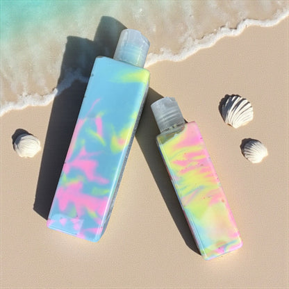 Two Bottles of Body Creme Lotion in the scent Tan lines. The bottles are clear and the lotion inside is swirls of yellow, pink, and blue. They are laying flat on a sandy beach with the ocean tide coming up around them. there are small white seashells scattered around the bottles. 