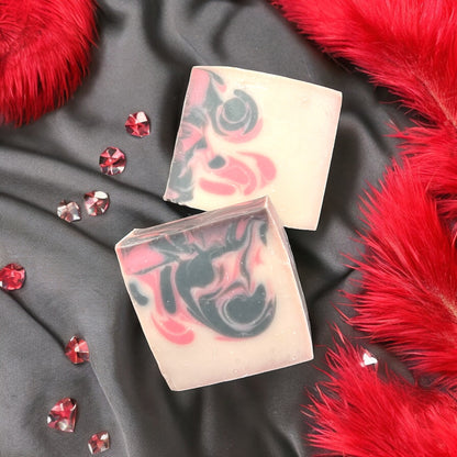 Two bars of swirled white, black and red pattern bar soap on a black satin textured background with red feathered accents and decorative red crystals scattered around.