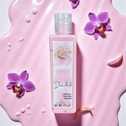 a bottle of body creme lotion in the scent drenched lays flat on a white surface with pink liquid surrounding it and purple orchids scattered around the bottle. 