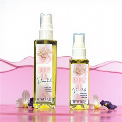 Two bottles of Drenched body oil, one 2 ounce and one 4 ounce bottle, with pink design label, against a white background with a liquid splash of pink behind it and purple and white flowers scattered around the bottles