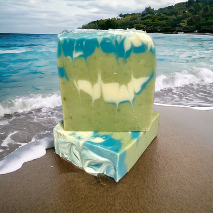 two bars of soap, one sitting flat while the other stands on top of it. both bars are green in color with shades or white and light blue marbled throughout the bars. both bars sit on a sandy beach with the tide coming in behind them. the ocean is in the background with a green lush mountain in the background