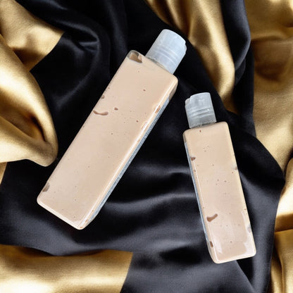 Two bottles of Dandy Moon body creme lotion in the scent cocoa butter cashmere. the bottles are clear and the lotion inside are a beige gold. the bottles are laying flat on a satin and silk fabric that is gold and black. 