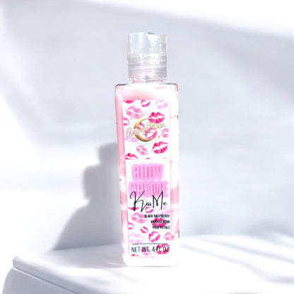 a bottle of body creme lotion in the scent kiss me. it is a clear bottle with reddish pink and white lotion inside. the label on the bottle is white with red and pink lips on it. the bottle stands on a white surface against a white background. 