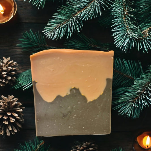 A bar of Dandy Moon Soapery's Aspen Woods soap is displayed against a rustic, woodsy surface, with pine branches, pine cones, and a warm candle. The soap has a layered design, with earthy tones resembling a  mountain silhouette beneath a light, natural beige sky. The simplicity and natural color palette evokes the serenity of winter forest landscape. 