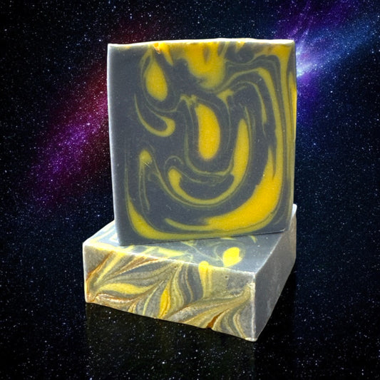 A bar of Dandy Moon Soapery's Astro mens Soap with a bold swirl design in dark navy blue and bright yellow. The bar sits atop another bar, to show the gold swirl design of the bar. Both bars are set against a galaxy and stars background. The contrasting colors and intricate marbling evoke a sense of energy and sophistication, making this handcrafted soap a luxurious choice. 