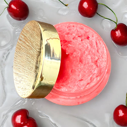 an open jar of cherry bomb foaming sugar scrub is seen from the top, showing inside the jar. the gold lid of the jar is resting on the side of the jar. the jar sits on a water rippled white surface. there are cherries all around the jar