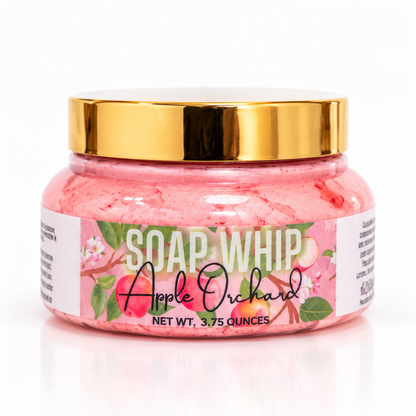 A jar of Soap Whip 'Apple Orchard'  with reddish whipped soap inside, a shiny gold lid, and a white label decorated with pink apples and green leaves, set against a clean white background.