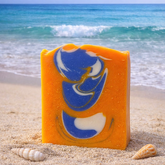 Bar of soap that is bright orange with a blue and white swirl design on a sandy beach with ocean waves in the background. there are two seashells scattered around the soap in the sand.