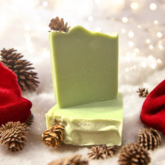 A bar of Dandy Moon Soapery's Cedar and Pine soap is displayed on a soft, snow like backdrop with festive elements, including pinecones and a hint of red fabric. The soap has a gentle green hue, capturing the natural essence of evergreen trees. It's simple earthy appearance embodies a refreshing, woodsy vibe, ideal for winter. This handcrafted soap offers a cozy forest inspired self care experience perfect for the holiday season. 