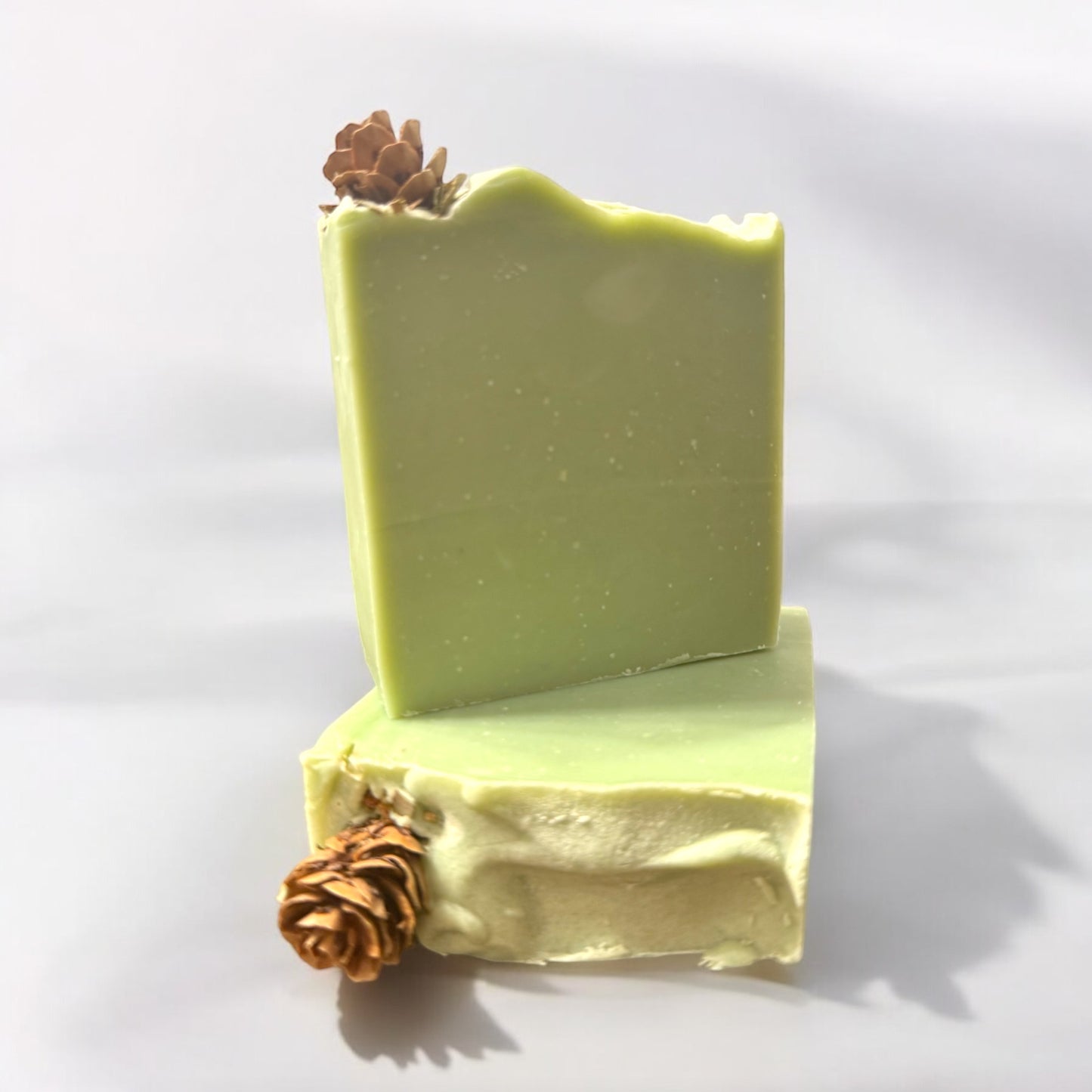 A bar of Dandy Moon Soapery's Cedar and Pine soap is displayed against a white backdrop on a white surface. The soap has a gentle green hue, capturing the natural essence of evergreen trees. It's simple earthy appearance embodies a refreshing, woodsy vibe, ideal for winter. This handcrafted soap offers a cozy forest inspired self care experience perfect for the holiday season. 