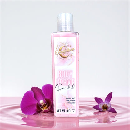 a bottle of dandy moon body creme lotion stands on a light pink rippled surface with purple orchids scattered around them. it stands against a white background. 