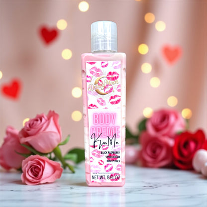 Bottle of 'Kiss Me' body creme lotion with pink and red lips on the label, in a clear bottle with pink and white lotion inside the bottle. the bottle stands on a white surface with hearts, twinkle lights, and red and pink roses scattered in the background. 