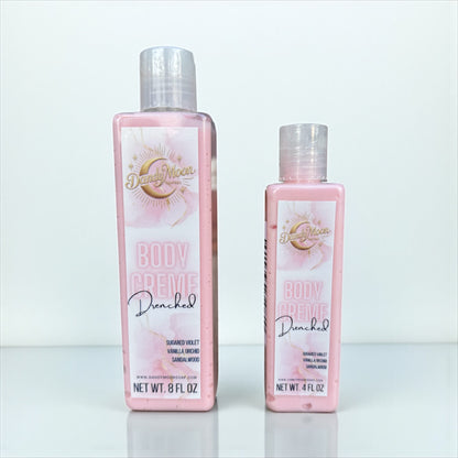 two bottles of dandy moon body creme lotion in the scent drenched stand on a white surface against a white background. the bottles are clear and the lotion inside is light pink. 