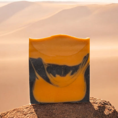 A bar of soap with a black and orange color design, placed against a backdrop of a desert landscape.