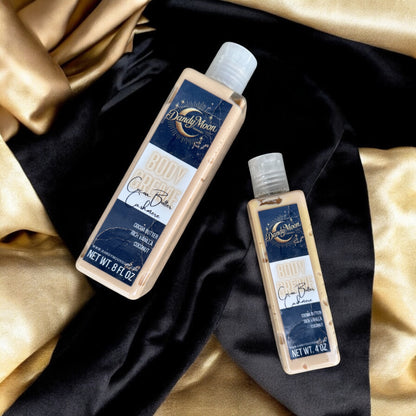 Two bottles of Dandy Moon body creme lotion in the scent cocoa butter cashmere. the bottles are clear and the lotion inside are a beige gold. the bottles lay flat on a satin gold and black surface. 
