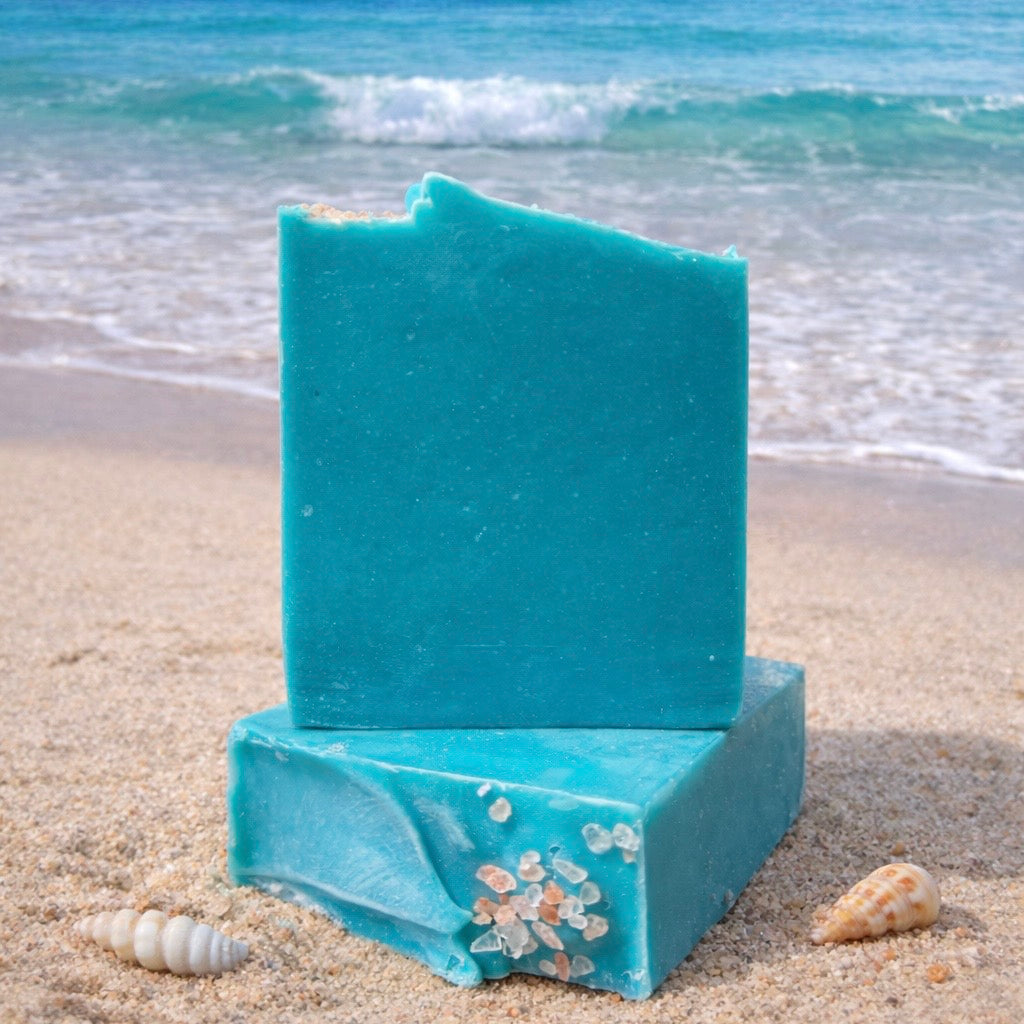 Two bars of blue soap, one is laying flat on its side to showcase the top design with himalayan sea salt top embeds, the other stands on top of that bar, with sea salt on a sandy beach with ocean waves in the background.