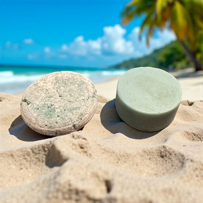 a light blue round shampoo bar and a light blue conditioner bar sit in the sand on a beach, begind them is a blurry backgound of the ocean and palm trees 