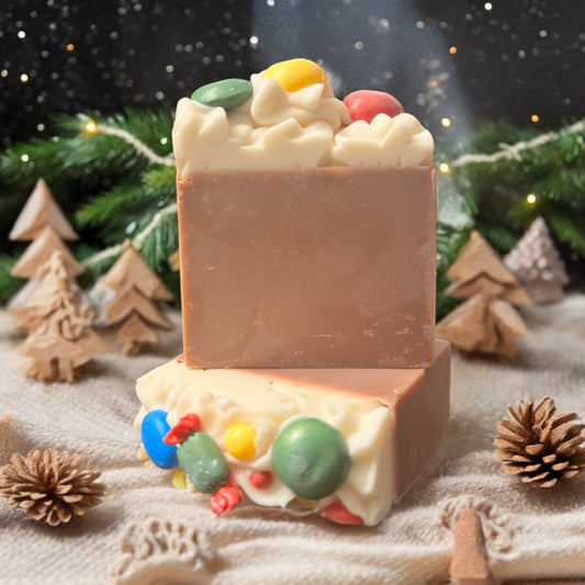 A bar of festive soap from Dandy Moon Soapery is displayed in a cozy winter setting, surrounded by miniature pine trees made of gingerbread, pine branches, and pine cones. The soap has a warm brown color with a decorative white top, adorned with colorful candy like soap embeds of yellow, green, red, and blue. The playful holiday inspired design evokes a joyful vibe, perfect for the holidays. 