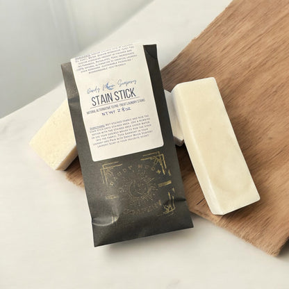 a black paper envelope with a logo on it that reads stain stick by dandy moon soapery. there is another stain stick to it and two others under neither the black envelope. all three bars are rectangular in shape and pale white. the envelope and stain sticks sit on a wooden accent that sits against a white backdrop. 