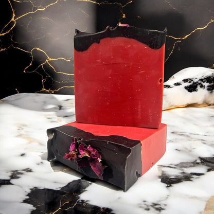 A photo of a two bars of handmade soap, one lying flat to show the top design with dried rose petals, and the other bar sitting on top of it, with a swirl of black activated charcoal on top, positioned on a marble surface, with a blurred image of a flower in the background.