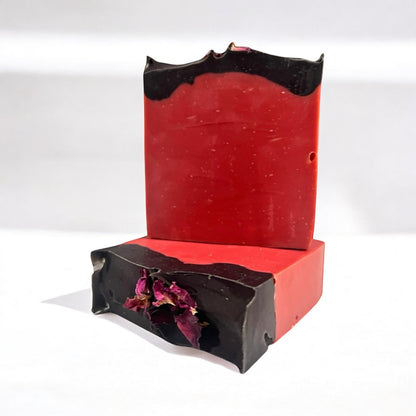 A photo of a two bars of handmade soap, one lying flat to show the top design with dried rose petals, and the other bar sitting on top of it, with a swirl of black activated charcoal on top. both sit on a white surface against a white background.