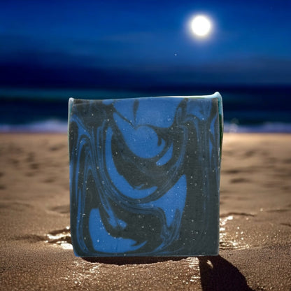 A bar of soap with black and blue swirls, placed on a sandy beach at night with the moon and ocean in the background.