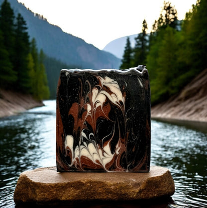 A bar of soap with swirls of black, brown, and white, placed on a rock with a scenic background of trees and mountains.