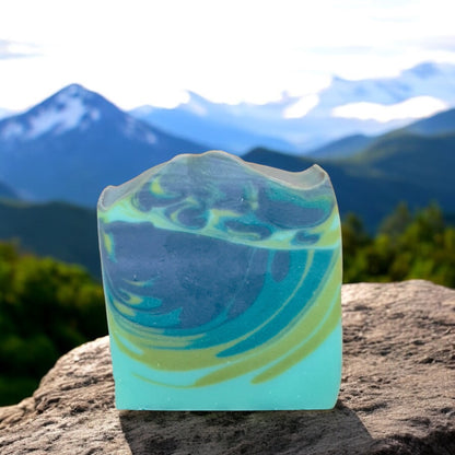 A bar of soap with light blue, green and darker blue swirls, placed on a natural rock with a mountainous background.