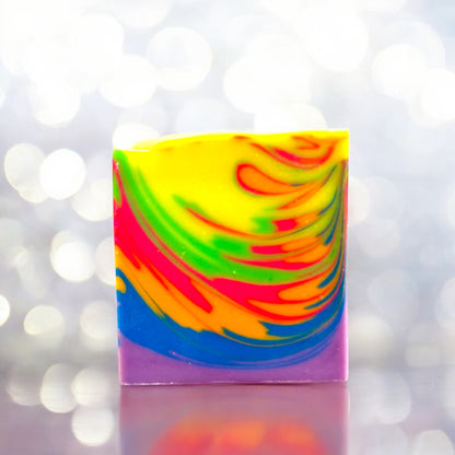 A multicolored bar of soap with a neon rainbow wave design, displayed against a sparkling background sitting on a reflective surface