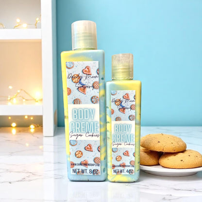 Two bottles of Dandy Moon Body Creme Lotion in the scent Sugar Cookies sit on a white marble countertop with a plate of sugar cookies beside them. The background is light blue and there is a white shelf to the left in the background that has twinkle lights draped on the shelf. The bottle of lotions are clear and the lotion inside them is a swirl of blue and yellow. 