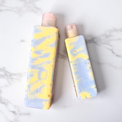 Two bottles of body creme lotion lay on their side to showcase the clear bottles with swirls of blue and yellow lotion inside them, laying flat on a marbled white surface