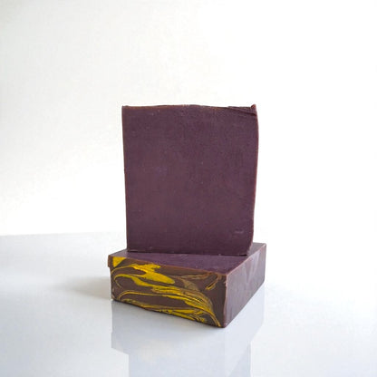 two Bars of soap, one sitting on top of the other. both bars are a deep rich purple, with the bottom bar showing its top design of golden swirls. both bars sit on a white surface against a white background