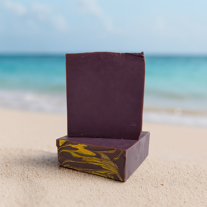 two Bars of soap, one sitting on top of the other. both bars are a deep rich purple, with the bottom bar showing its top design of golden swirls. both bars sit on a sandy beach with the ocean blurred in the background