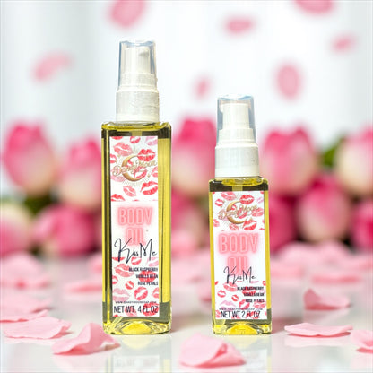 Two bottles of body oil, one is 2 ounces and one is 4 ounces, with labels that have a lips design, on a white surface with pink rose petals scattered around the bottles, with a blurred background or pink rose bouquets.
