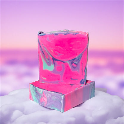 two Colorful soap bars with a gradient swirl of pink, purple and light blue. one bar is laying flat while the other is standing on top of it. the bars sit on a light pink cloud with a blurred cloudy pink and purple effect in the background