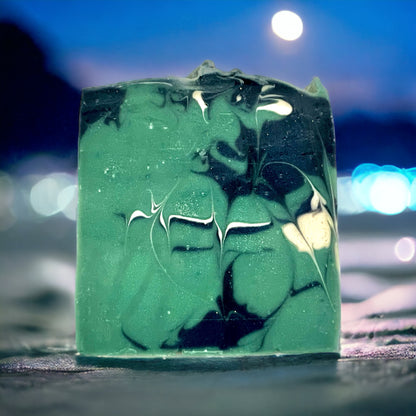 A bar of handmade soap with a green and white marbled design, topped with a dark green layer, set against a blurred moonlit background.