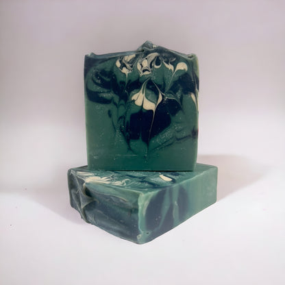 Two bars of handmade soap with a green and white marbled design. One bar lays flat to show the top design while the other sits on top of it. both bars sit on a white surface against a white background.
