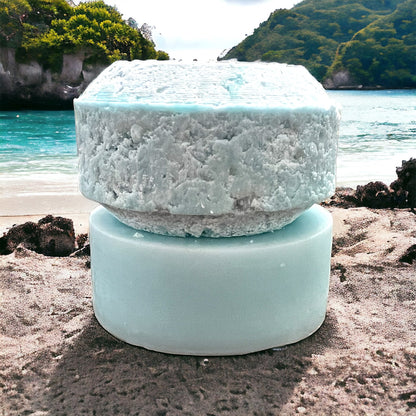 a stack of two bars. the one on the bottom is a blue hue and is round and smooth, with another puck shaped round blue hue bar on top of it. the one on top is the shampoo, the one on bottom is the conditioner. both bars sit on a sandy surface, with a beach and green lush mountains in the background.