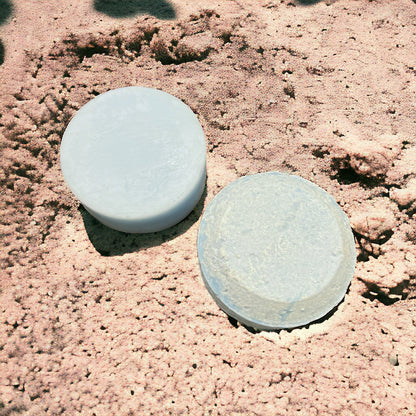two blue bars sit on a sandy surface. one blue bar is on the left and is smooth. its the conditioner bar. the one on the right is the shampoo bar and it is also a blue hue color and is round and puck shaped. 