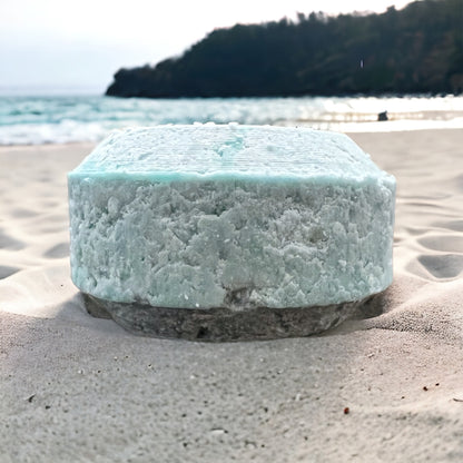 a light blue puck shaped shampoo bar sits in the sand on a beach with a blurred ocean and mountain background.